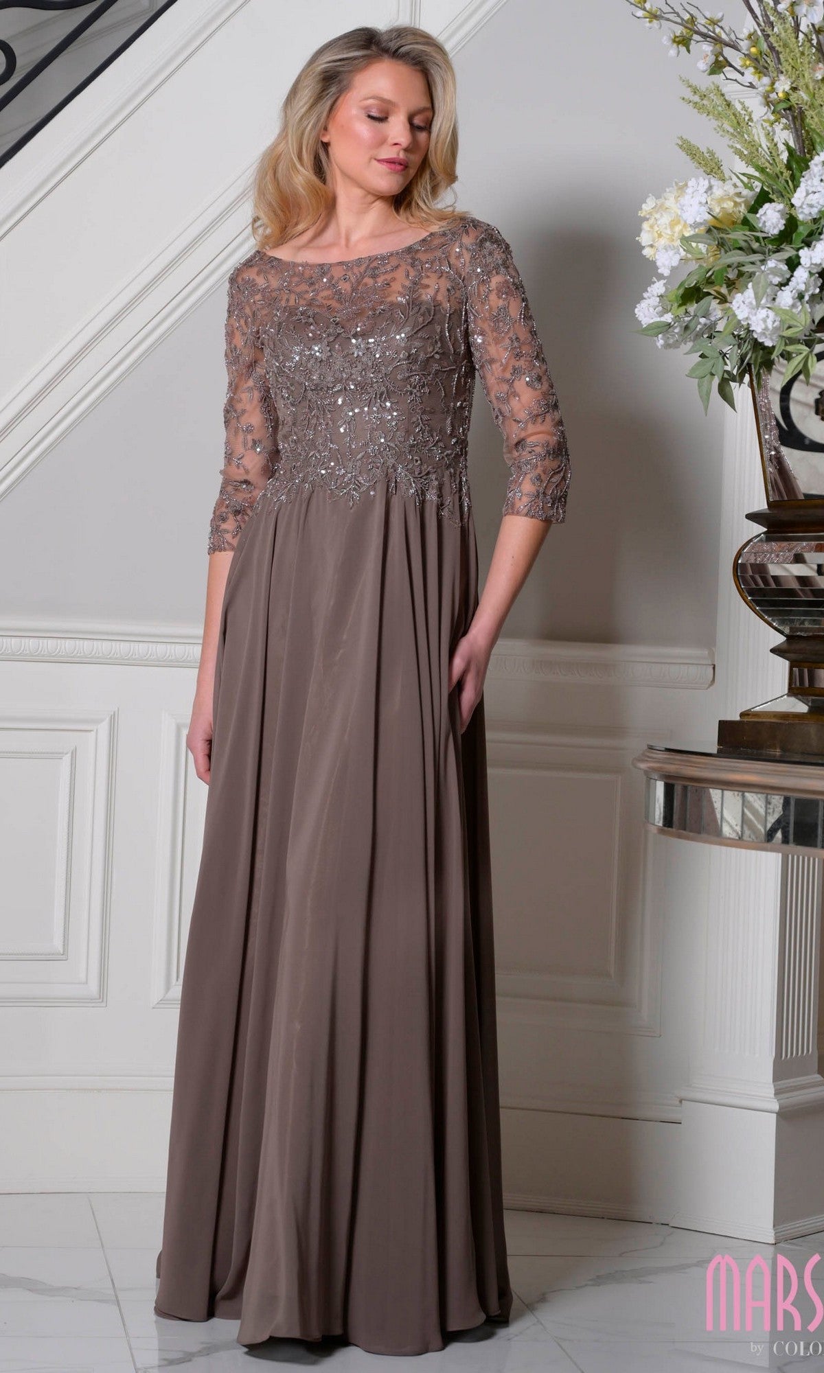 A woman in the Marsoni M340SL long formal dress in dark taupe, featuring a beaded-embroidered bodice, sequin accents, three-quarter sleeves, and an elegant A-line chiffon skirt, stands indoors by a staircase and floral arrangement.